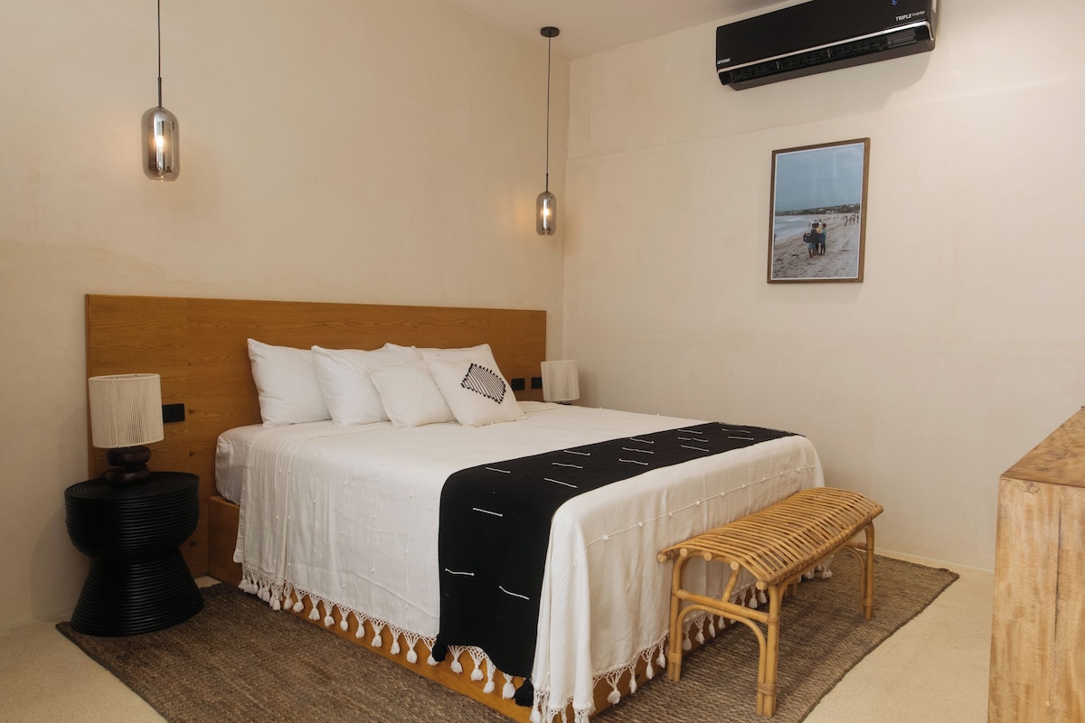 Un dormitorio con cama, mesita de noche, lámpara, unidad de aire acondicionado y un cuadro en la pared.