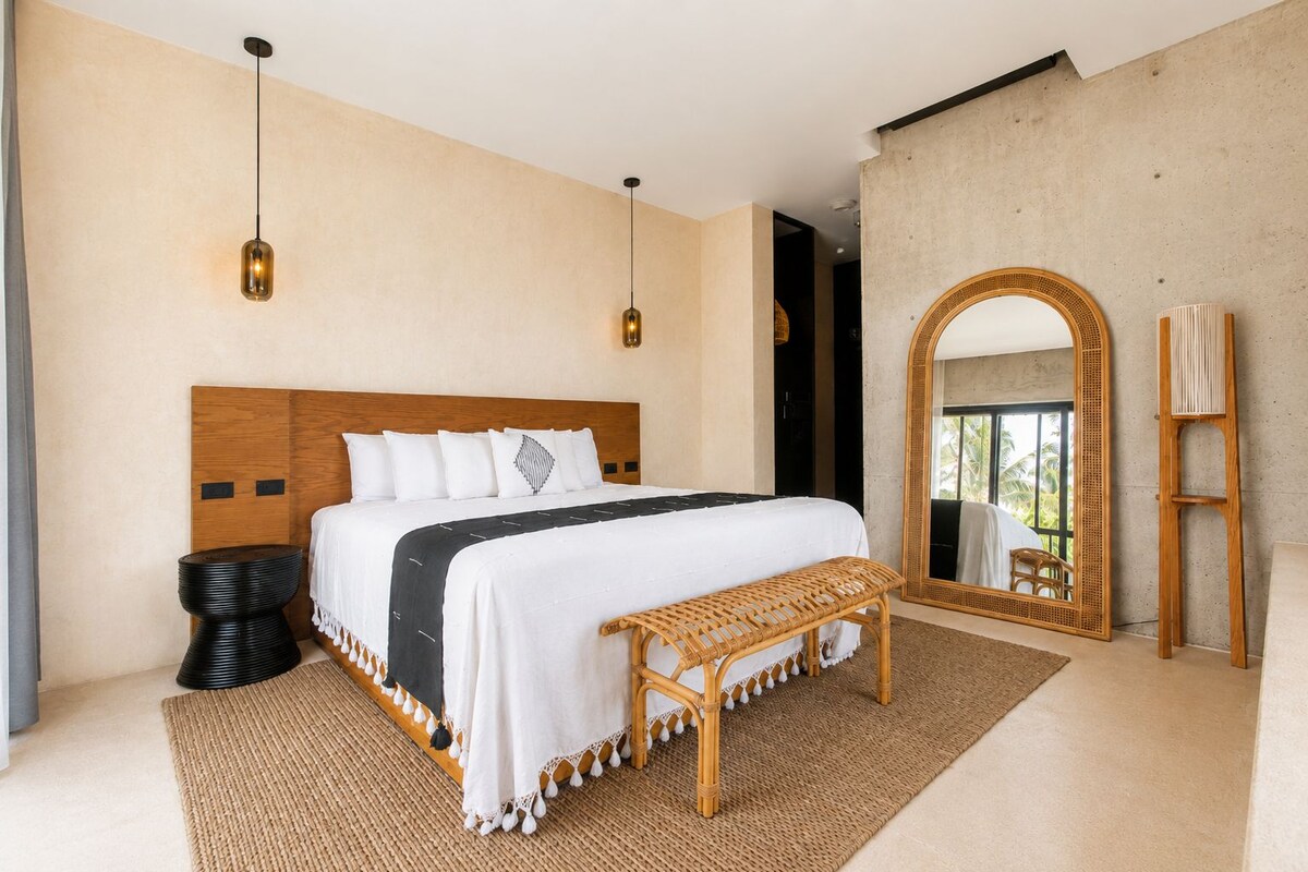 Un dormitorio con una cama grande, dos lámparas colgantes, un cabecero de madera, un banco, un espejo grande y vistas al exterior.