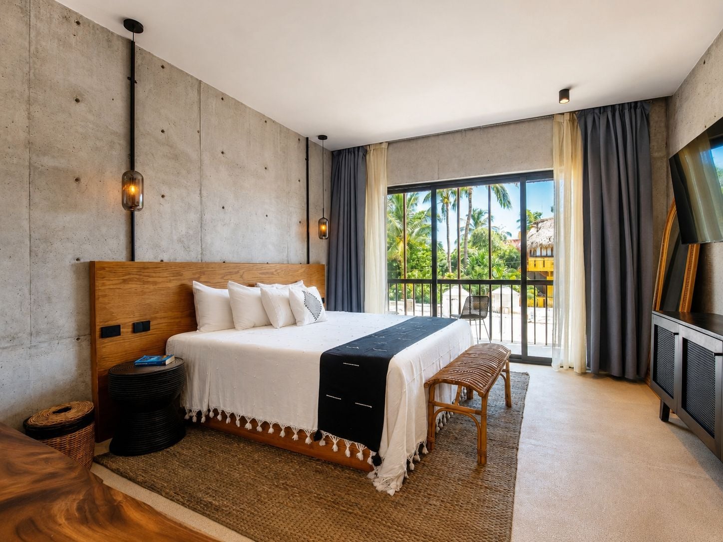 Habitación de hotel moderna con una cama grande, un balcón con vista a palmeras y un televisor a la derecha.