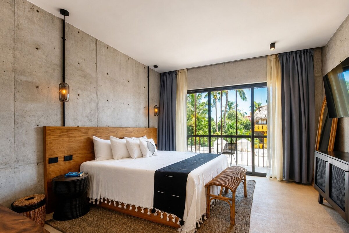 Habitación de hotel moderna con una cama grande, un balcón con vista a palmeras y un televisor a la derecha.