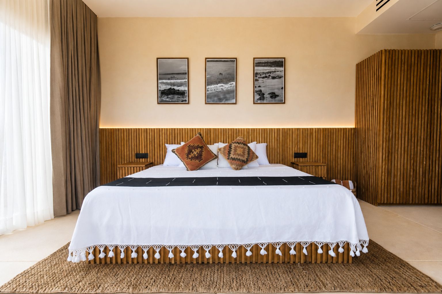 A hotel room with a large bed, a wooden headboard, a woven rug, and three framed pictures on the wall.