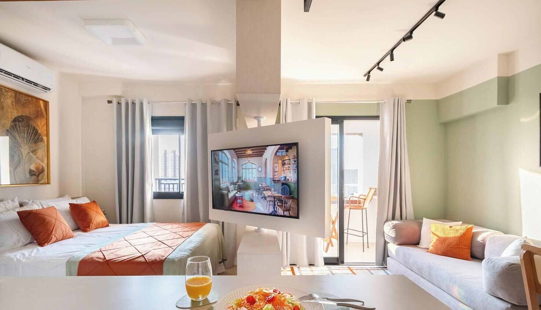 Habitación de hotel con cama, televisión, mesa de comedor y vaso de jugo de naranja, y vista a la ciudad.