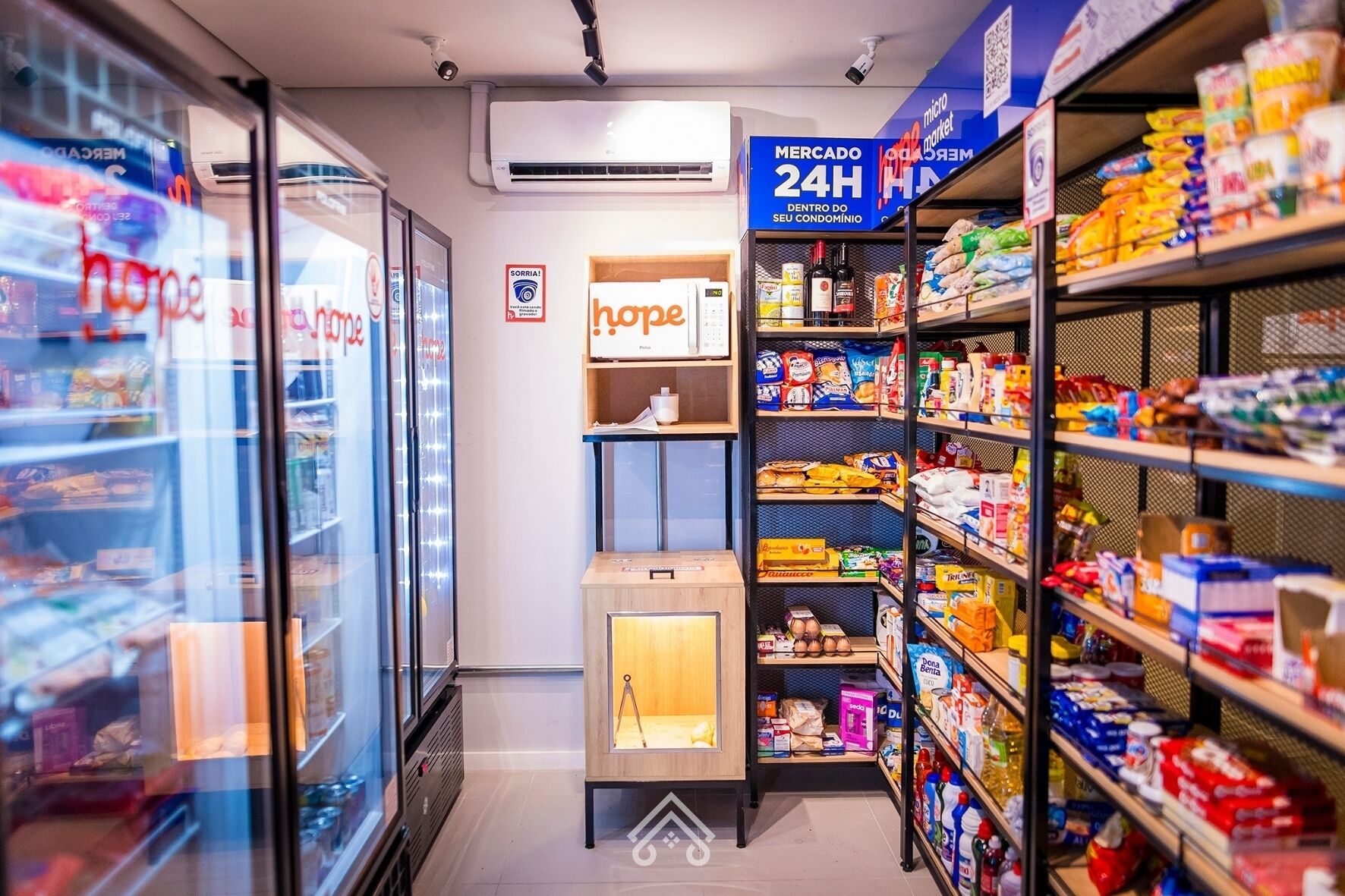 Interior de um supermercado com seções refrigeradas, prateleiras repletas de diversos produtos e uma placa indicando um mercado 24 horas dentro de um condomínio.