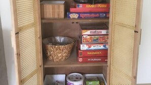 A wooden cabinet with open doors, containing board games and a woven basket. - Bendalong  Noordwijk (Noordwijk)