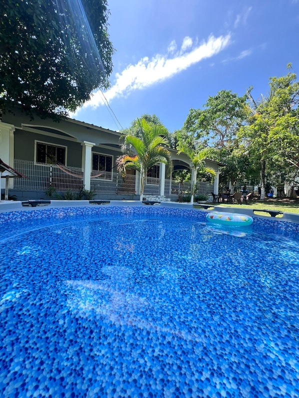 Une piscine dont le fond est recouvert de mosaïques bleues, entourée d’un espace délimité par une clôture, avec des arbres et un bâtiment en arrière-plan.