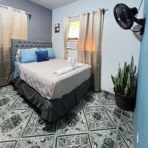 Une chambre à coucher avec un lit, un ventilateur de chevet, une plante en pot et des carreaux de sol à motifs.