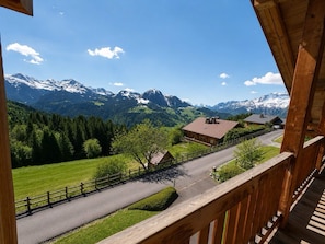 Ein hölzerner Balkon mit Blick auf eine Bergstraße, mit Häusern und schneebedeckten Gipfeln in der Ferne.