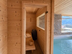 A wooden sauna room with a bench, a stone wall, and a window overlooking a pool. - Luxury Chalet "La Framboiseille" with Pool and Sauna (Manigod)