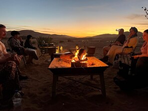 A group of people seated around a campfire in a desert setting with a view of the sunset.