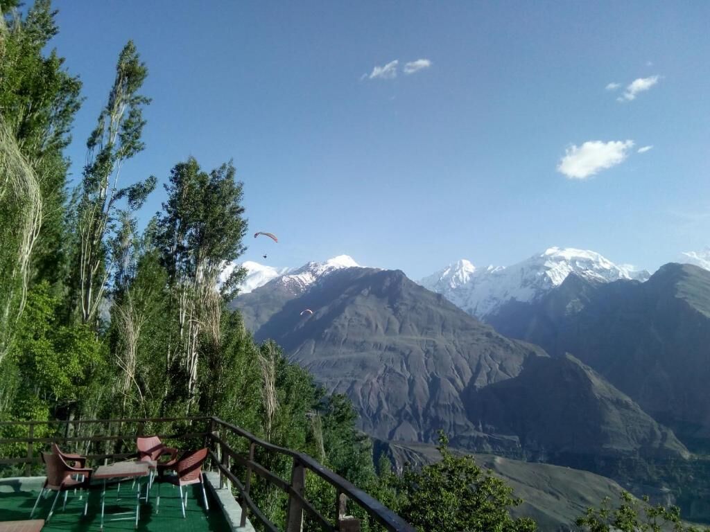 A mountainous landscape with a clear sky, a chair and table set up on a platform, and a person paragliding in the distance.