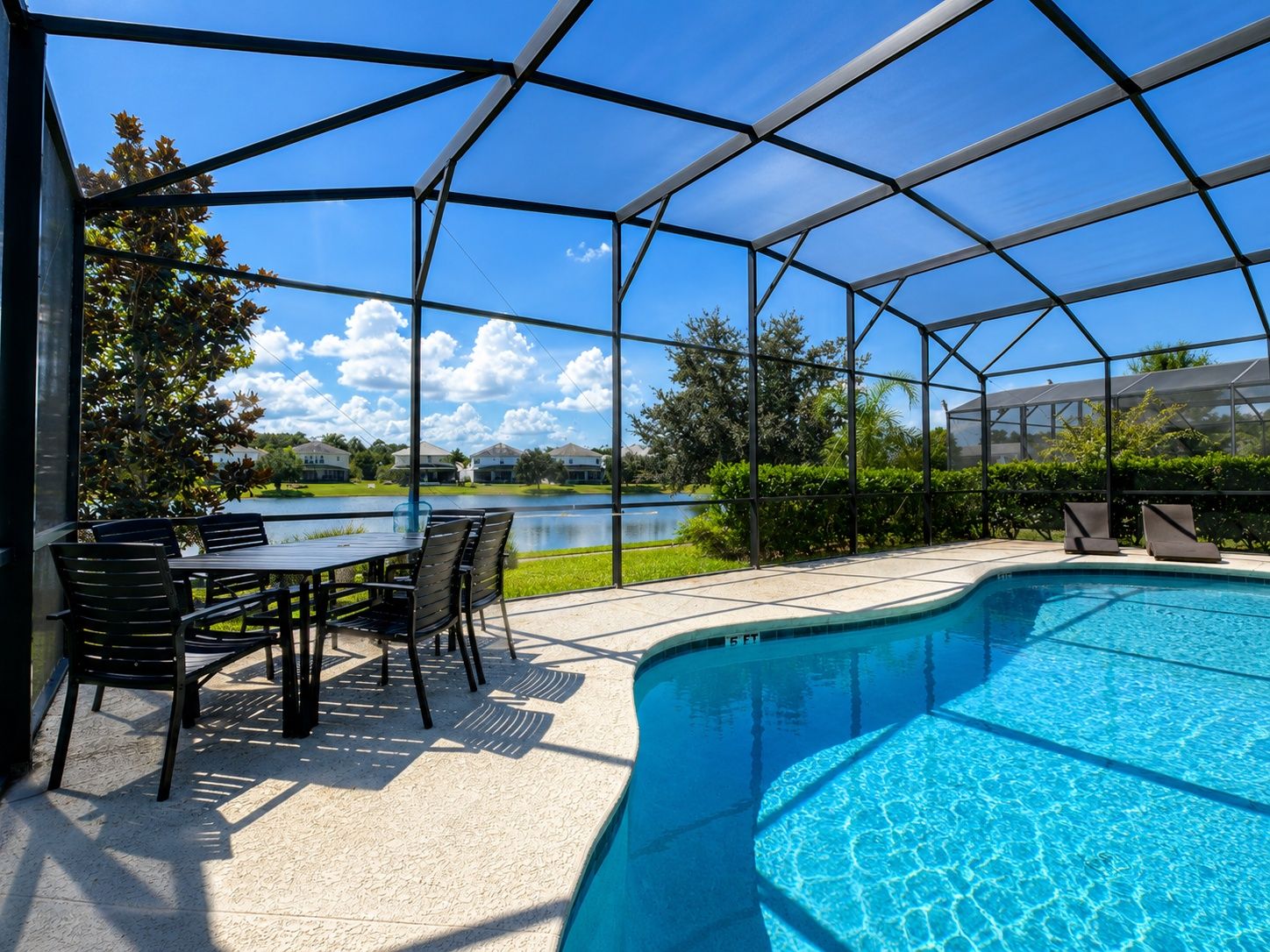 Piscina exterior cubierta con zona de patio, una mesa y sillas, y vistas a un lago y casas al fondo.