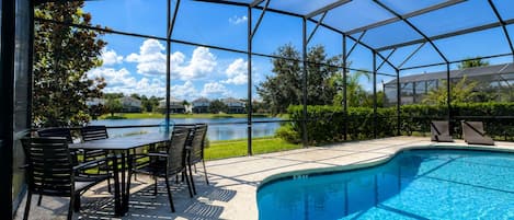Piscina exterior cubierta con zona de patio, una mesa y sillas, y vistas a un lago y casas al fondo.