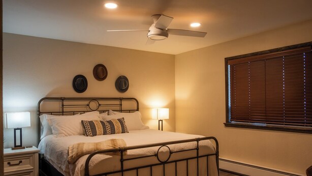 Quarto com cama, abajures, ventilador de teto e janela com cortinas.