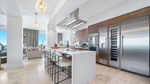 A modern kitchen with stainless steel appliances, a central island with bar stools, and a view of the city. - Beach Club Avalon Penthouse #1 - Luxury Beachfront 4BR, Views, Pool, Hot Tub (Gulf Shores)