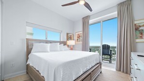 A hotel room with a large bed, a ceiling fan, a window with a view, and a balcony with a chair. - Beach Club Avalon Penthouse #1 - Luxury Beachfront 4BR, Views, Pool, Hot Tub (Gulf Shores)