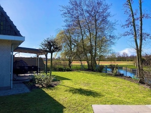 A covered outdoor seating area with a table and chairs, a grassy lawn, and a body of water in the background. - Maralong Noordwijk (Noordwijk)
