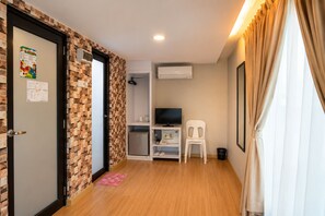 A hotel room with a brick-patterned wall, a small kitchenette, a television, a white plastic chair, and a window with curtains.