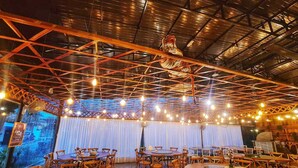 An indoor dining area with multiple round tables and chairs, a high ceiling with exposed beams, and string lights.