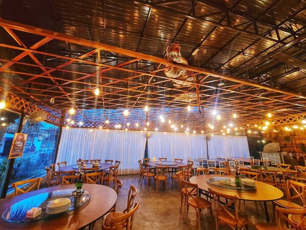 An indoor dining area with multiple round tables and chairs, a high ceiling with exposed beams, and string lights.