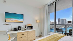 Ein Hotelzimmer mit großem Fenster, einem Bett, einem Schreibtisch mit Computer, einer Kaffeemaschine und einem Strandbild.