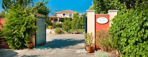 A gated entrance with a sign, a paved driveway, and a building with a red roof in the background. - Lefkas Blue Residence Two-bedroom Suite near to the town and close to the beach. (Lefkada)
