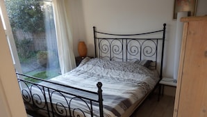 Uma cama com estrutura de metal, uma mesa de cabeceira e uma janela com cortinas.