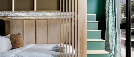 Una cama litera con cabecera de madera, una cama individual con almohada y una escalera frente a una pared verde.