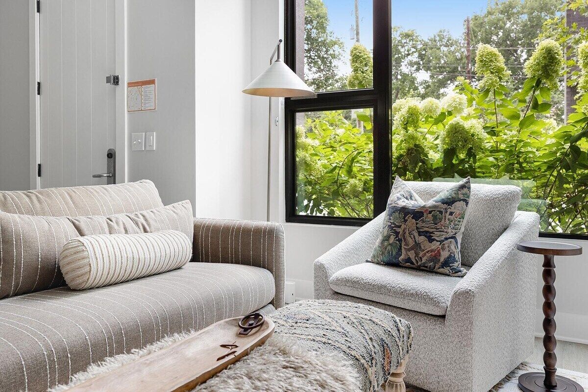Un salon avec un canapé et un fauteuil, une table basse en bois, une lampe et une vue sur la verdure à l’extérieur.