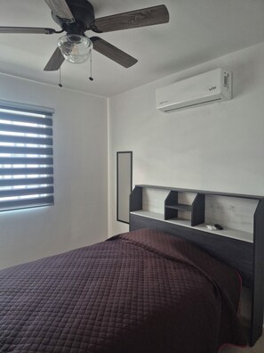 Ein Schlafzimmer mit einem Bett, einem Deckenventilator, einer Klimaanlage, einem Fenster mit Jalousien und einem Einbauschränkchen.