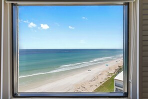 A beach view through a window with a clear sky and calm sea. - 🏖️ Seascape Towers 1025 | Direct Oceanfront Condo | Balcony + Pool Access (New Smyrna Beach)