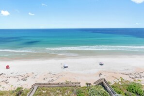 A beach with a wooden boardwalk leading to a sandy area with a few beach umbrellas and a red lifeguard chair. - 🏖️ Seascape Towers 1025 | Direct Oceanfront Condo | Balcony + Pool Access (New Smyrna Beach)