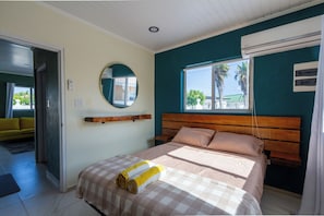 Ein Schlafzimmer mit einem Bett, einem Nachttisch, einem Spiegel und einem Fenster mit Blick auf Palmen.