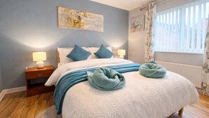 Ein Schlafzimmer mit einem Bett, Nachttischlampen, einem Beistelltisch, einem Fenster mit Vorhängen und einem Wandbild.