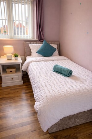 A neatly made bed with a white comforter and a rolled towel, a bedside table with a lamp and a small plant, and a window with curtains. - Huge 4BR Detached Family Home Near Derby City Centre (Derby)
