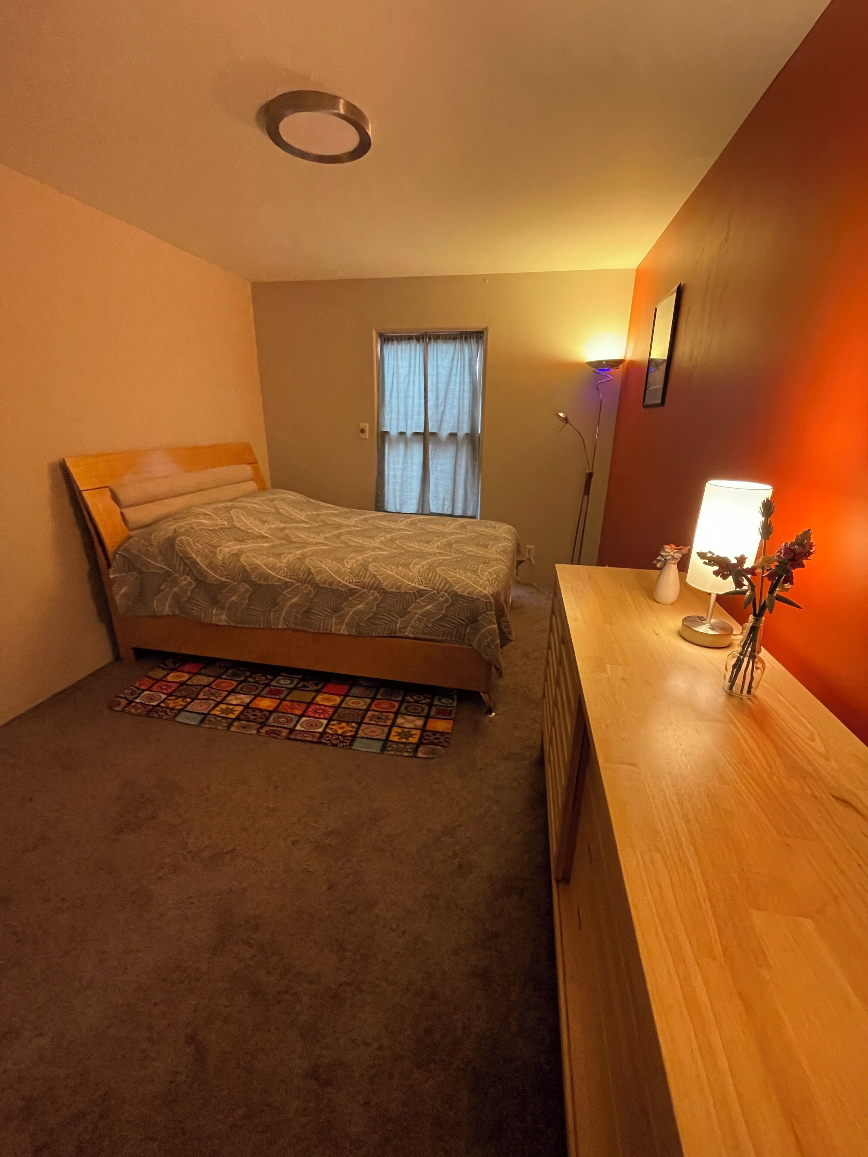 A bedroom with a bed, a bedside table with a lamp, a window with curtains, and a wall-mounted light fixture.