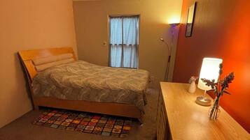 A bedroom with a bed, a bedside table with a lamp, a window with curtains, and a wall-mounted light fixture.