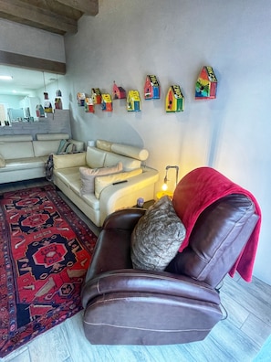 Una sala de estar con un sofá, un plaid rojo, un gato, un tapete estampado y una pared decorada con casas de pájaros de colores.