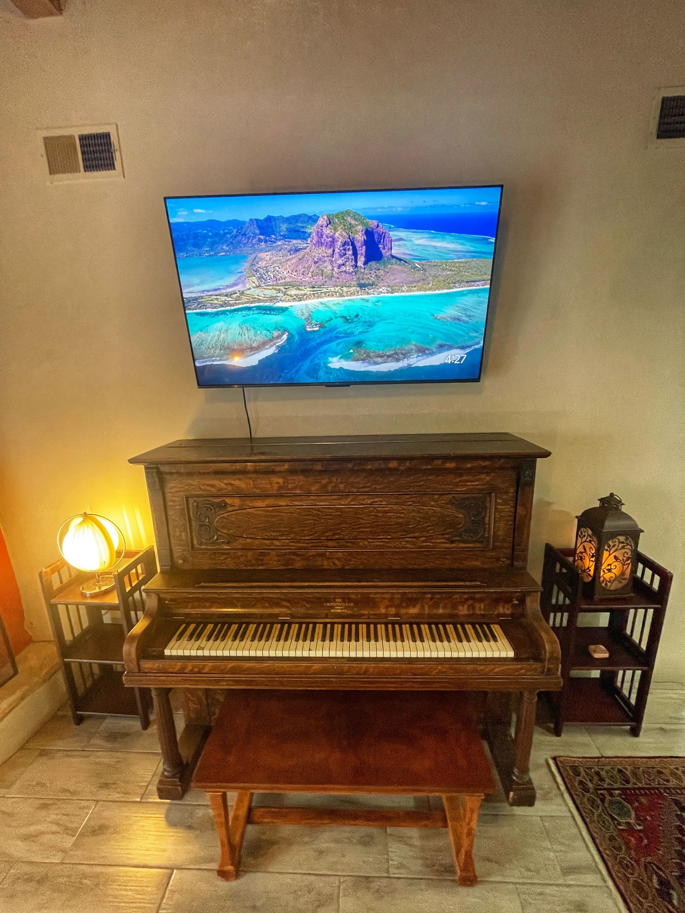 A wooden piano with a bench, a television mounted on the wall, and two wall-mounted lamps.