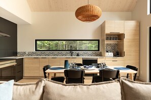 A modern kitchen with a dining area, featuring a large window, wooden cabinets, and a hanging light fixture. - Pamyland : A chic nature getaway by the water (Saint-Alphonse-Rodriguez)