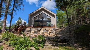 A modern cabin with a large glass window, wooden deck, and outdoor seating. - Pamyland : A chic nature getaway by the water (Saint-Alphonse-Rodriguez)