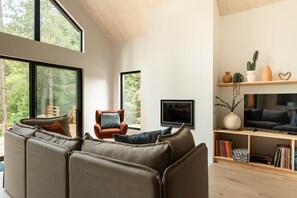 A modern living room with a large sectional sofa, a single armchair, a flat-screen TV, and a bookshelf with decorative items. - Pamyland : A chic nature getaway by the water (Saint-Alphonse-Rodriguez)