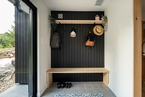 Un petit espace de véranda fermé, comprenant un banc, une étagère fixée au mur avec une plante, et un mur sur lequel sont accrochés un sac et deux chapeaux.