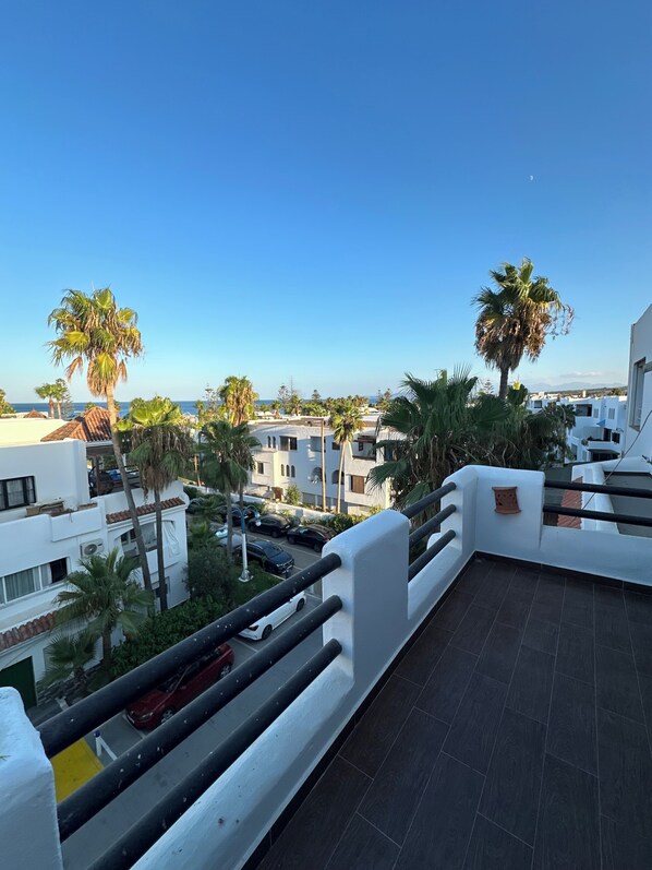 A balcony with a view of the sea, palm trees, and a street with cars.
