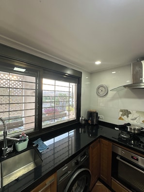 A kitchen with a stainless steel sink, a washing machine, a stove, and a window with a view of greenery. - Mediterranean Getaway (Fnideq)
