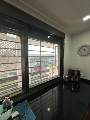 A kitchen with a black countertop, a clock on the wall, and a window with a view of greenery and buildings. - Mediterranean Getaway (Fnideq)