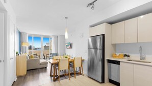 A modern kitchen with a stainless steel refrigerator, a dining area with wooden chairs, and a view of the city through large windows. - Sunset Views 2BR - 2BTH w/ Bay View Fitness Center (Hallandale Beach)
