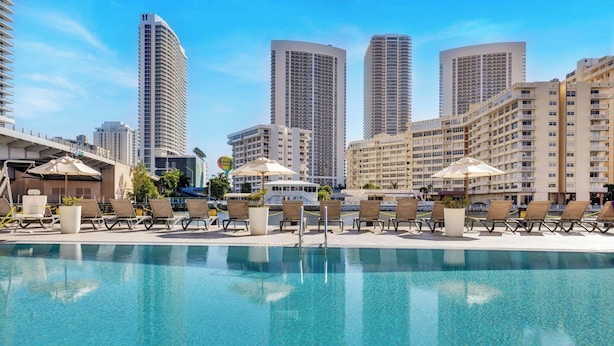 A poolside area with lounge chairs and umbrellas, surrounded by high-rise buildings. - Sunset Views 2BR - 2BTH w/ Bay View Fitness Center (Hallandale Beach)
