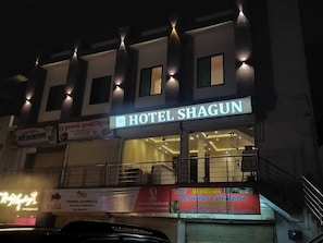 A hotel named 'HOTEL SHAGUN' with a signboard, balconies, and a closed shutter on the ground floor.