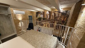 Un dormitorio con cama, una silla, un balcón con barandilla y vistas a un patio interior con arcos y plantas.