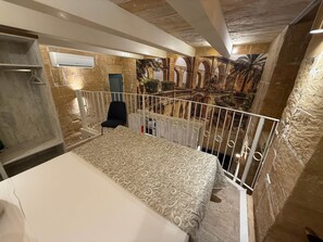 Ein Schlafzimmer mit Bett, Sessel, einem Balkon mit Geländer und Blick auf einen Innenhof mit Bögen und Pflanzen.